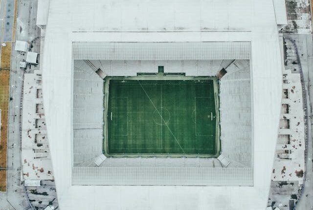 Vista aérea de um estádio de futebol vazio, com arquibancadas ao redor do campo verde e estrutura moderna vista de cima.